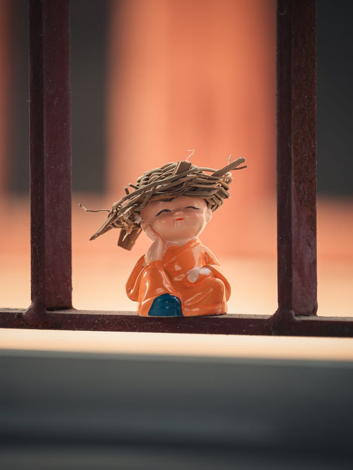 A cute figurine wearing an orange robe and a thatched hat, sitting on a windowsill.